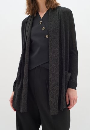 Svart, glittrig cardigan med öppen framdel, texturerad tyg, långa ärmar och två framfickor, lager-på-lager över en svart topp med knappar.