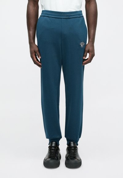 Zelenomodré teplákové nohavice z mäkkého materiálu s elastickým pásom a malým logom leva na ľavej stehne. Kombinované s čiernymi teniskami na šnurovanie.