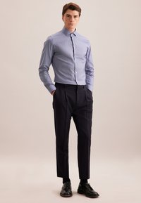 Man in een lichtblauw knoopshirt, zwarte maatpakbroek, zwarte sokken en zwarte nette schoenen, staande met de handen in de zakken.