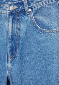 Gros plan sur un jean en denim bleu montrant la poche avant, la ceinture avec bouton, la boucle de ceinture et les détails de couture orange visibles.