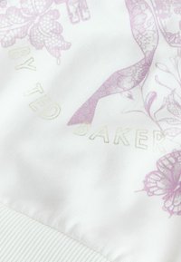 Valkoinen collegepaita, jossa vaaleanpunaisia kukka- ja perhoskuvioita, ja kohopainettu "by Ted Baker" -teksti kullansävyisellä kirjailulla. Pinta on strukturoitu ja helmassa resorikuvio.