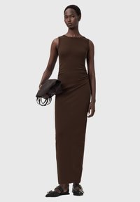 Brązowa sukienka maxi bez rękawów z elastycznego materiału, z prostą, dopasowaną sylwetką i bocznym rozcięciem. Modelka trzyma ciemną kopertówkę.