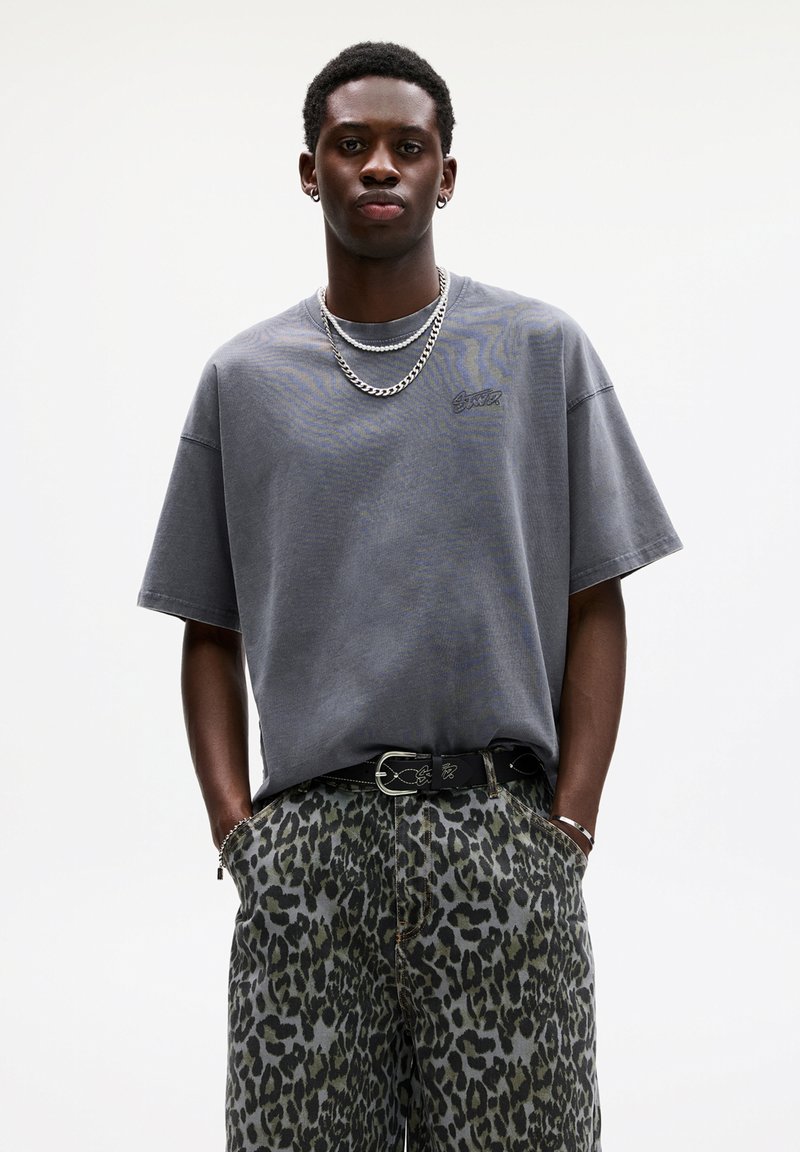 Grå oversized t-shirt med korte ærmer, der har et lille logo. Matchet med bukser i grøn og sort leopardprint og et sort bælte.