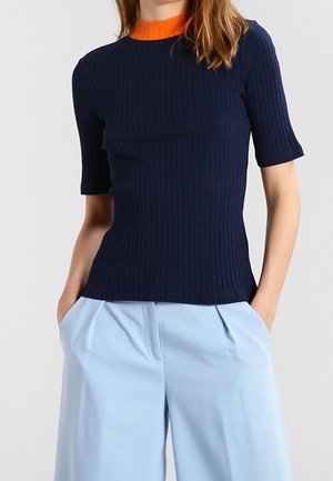 Persoon die een marineblauw geribd kort mouw shirt draagt met een oranje kraag en lichtblauwe wijde broekspijpen met handen in de zakken.