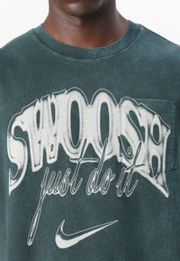 T-shirt in cotone verde scuro con una grande grafica bianca recante la scritta "SWOOOSH just do it" e una piccola tasca sul lato sinistro del petto. Presente il logo Nike.