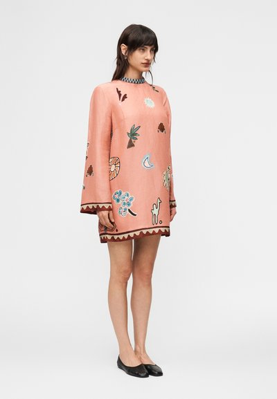 Γυναίκα που φοράει ένα φαρδύ, ροζ φόρεμα με μακριά μανίκια και πολύχρωμα αφηρημένα και φυσικά μοτίβα, συνδυασμένο με μαύρα μπαλαρίνες.