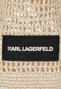 Tkanična torba od slame u svijetloj bež boji s crnom pravokutnom etiketom s bijelim natpisom "KARL LAGERFELD" u središtu.
