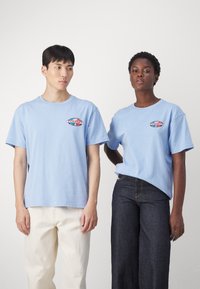 Camisetas de algodón de color azul claro con logo impreso en el pecho, corte relajado, mangas cortas; un modelo con pantalones crema y otro con vaqueros oscuros.