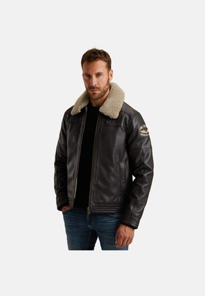 Veste en cuir marron avec un col en shearling beige, fermeture éclair à l'avant, poches latérales et un patch sur la manche gauche. Portée sur une chemise noire.