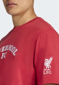 Detailný záber osoby s červeným tričkom Liverpool FC, na rukáve s bielym logom vtáka Liverpool a textom "LIVERPOOL FC" na hrudi.