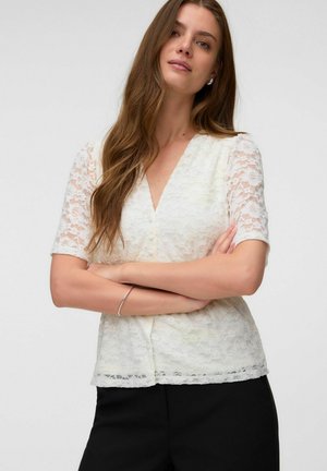 Frau mit langen braunen Haaren, die eine weiße Spitzenbluse mit kurzen Ärmeln und schwarze Hosen trägt, mit verschränkten Armen, stehend vor einem schlichten Hintergrund.
