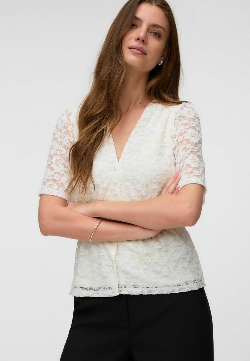 Femme aux longs cheveux bruns portant un chemisier blanc en dentelle à manches courtes et un pantalon noir, bras croisés, debout devant un fond uni.