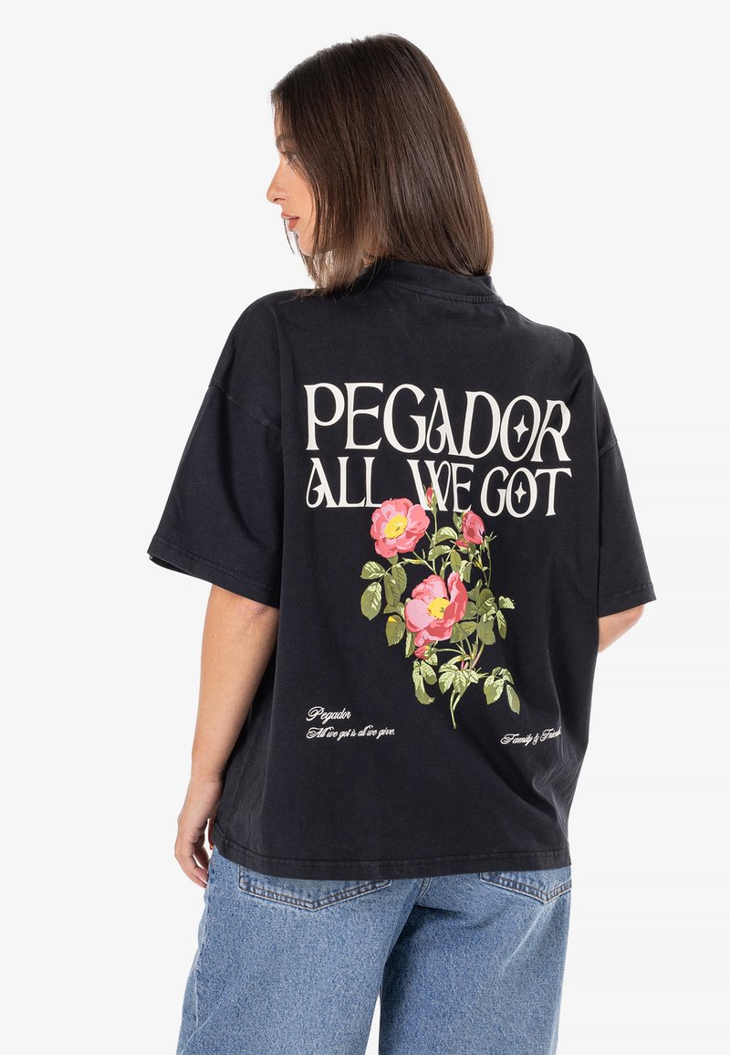 Čierne voľné tričko s kvetinovým dizajnom v ružovej a zelenej farbe na chrbte, s textom "PEGADOR ALL WE GOT" v krémovej farbe. Vyrobené z bavlny.