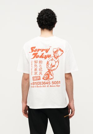 Bărbat cu păr creț purtând un tricou alb cu grafic de bomboană portocalie și detalii de contact ale magazinului Tokyo pe spate, pantaloni negri.