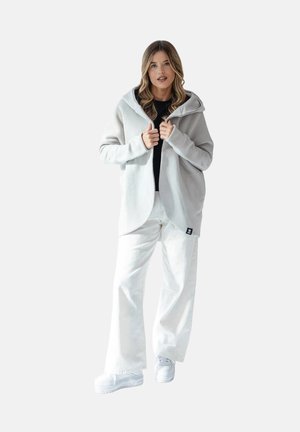 Kobieta w jasnoszarej bluzie z kapturem, czarnym topie, białych szerokich spodniach i białych trampkach, stojąca na jednolitym tle.