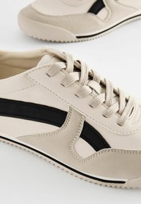 Beige nahkakengät mustilla yksityiskohdilla, tekstuuripaneeleilla ja beigeillä nauhoilla. Varustettu kumipohjalla, jossa on zigzag-kuvio.