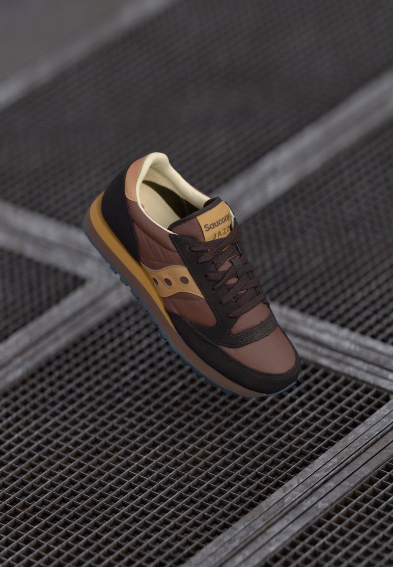 Brune sneakers med sorte detaljer, gul merke-logo, teksturert overdel og gummisåle. Har klassisk design med snøring.