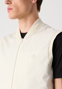 Beige īsrokas bomber jakas ar augstu apkakli, gludu audumu un viegli iespiestu logo uz krūšu daļas. Melna t-krekla redzama zem jakas.