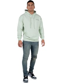Felpa verde chiaro con tasca anteriore a soffietto e cordini, abbinata a jeans blu slim fit con strappi e testo stampato, e sneakers bianche e nere.