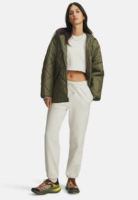 Young woman standing, wearing an olive green quilted jacket, white cropped sweatshirt, white sweatpants, and colorful athletic shoes.