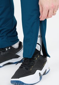 Turkosa sportbyxor med siddragkedja, i kombination med svarta sneakers med vita och blå detaljer, samt synliga vita strumpor.