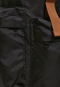 Tissu en nylon noir présentant plusieurs poches, fermetures éclair et une sangle ajustable en brun. Présente une surface texturée et des coutures renforcées.