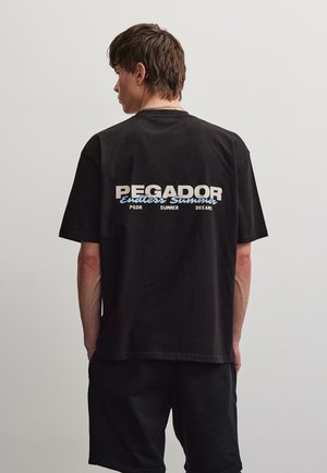 Fiatal férfi rövid barna hajjal, fekete pólót visel, amelyen a hátoldalon a „PEGADOR Endless Summer PGDR SUMMER DREAMS” felirat olvasható.