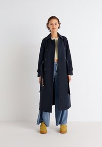 Navy trench coat with double-breasted buttons, long sleeves, and belt. Paired with light jeans and yellow platform boots. Smooth texture.