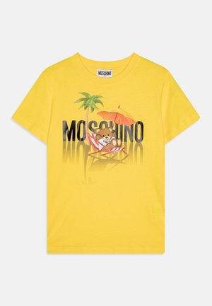 T-shirt jaune Moschino avec un ours en peluche se prélassant sur une chaise de plage rayée sous un parasol orange et un palmier.