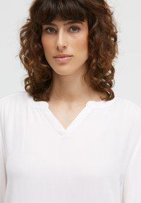 Frau mit lockigem braunem Haar und blauen Augen, die ein weißes V-Ausschnitt-Oberteil vor einem schlichten Hintergrund trägt.