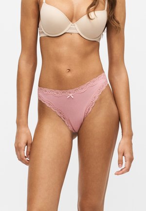 Femme portant un soutien-gorge beige clair et un dessous rose orné de dentelle, debout devant un fond blanc uni.