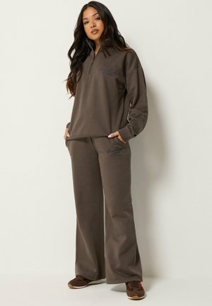 Femme portant un sweat-shirt marron oversize et un pantalon à jambes larges avec poches, debout contre un mur clair uni, les mains dans les poches.