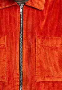 Giacca in corda rossa con chiusura frontale a zip, due ampie tasche a toppa e un design a coste testurizzato.
