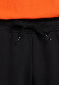 Pantaloni sportivi neri con cordino in vita, dotati di una cintura testurizzata e di una piccola etichetta. Una maglietta arancione è visibile sopra la cintura.