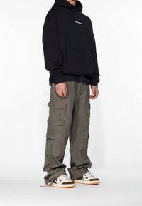 Zwarte capuchontrui met een voorzak, gecombineerd met olijfgroene cargobroek met meerdere zakken. Schoeisel bestaat uit bruine en crèmekleurige sneakers.