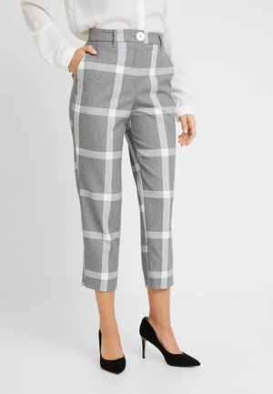 Femme portant un pantalon court gris avec un grand motif à carreaux blanc, une blouse blanche et des escarpins noirs à talons hauts, debout avec une main dans la poche.