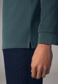 Grøn bomulds langærmet top med tekstureret stof, der har slidser i siderne ved kanten og tætsiddende manchetter. Sættes sammen med mørke denim jeans.