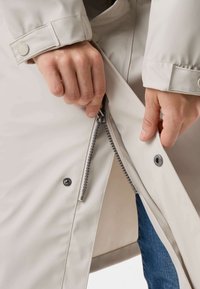 Des mains fermant une veste beige clair à boutons-pression, dévoilant partiellement un jean bleu en dessous.