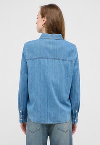 Jasnoniebieska koszula dżinsowa z długimi rękawami, zapinanymi mankietami oraz klasycznym kołnierzykiem; posiada karczek z tyłu i subtelną pionową fakturę.