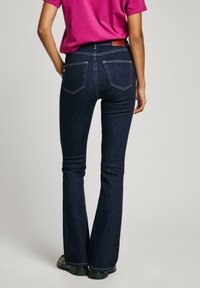 Jeans de mezclilla de tiro acampanado en azul oscuro con cuatro bolsillos, costuras decorativas y un parche de cuero marrón en la parte posterior. Ajustados en la pierna.
