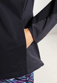 Main d’une personne dans la poche d’une veste noire, montrant la texture du tissu de la veste et des leggings colorés à motifs en dessous.