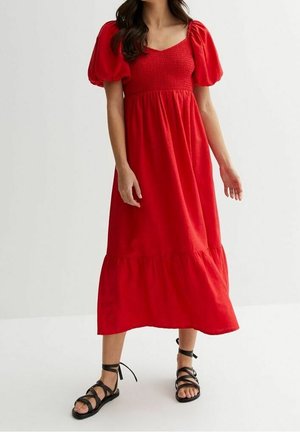 Vrouw draagt een rode midi-jurk met pofmouwen en zwarte platte sandaaltjes met veter, staand tegen een effen lichte achtergrond.