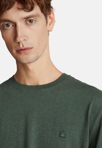 Zelené tričko s krátkým rukávem vyrobené z měkké tkaniny. Má kulatý výstřih a malou vyšívanou námořní potvoru na hrudi.