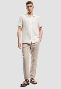 Korte mouw, crèmekleurig shirt met een textuurpatroon en knopen aan de voorkant, gecombineerd met lichtbeige broeken en zwarte sandalen.