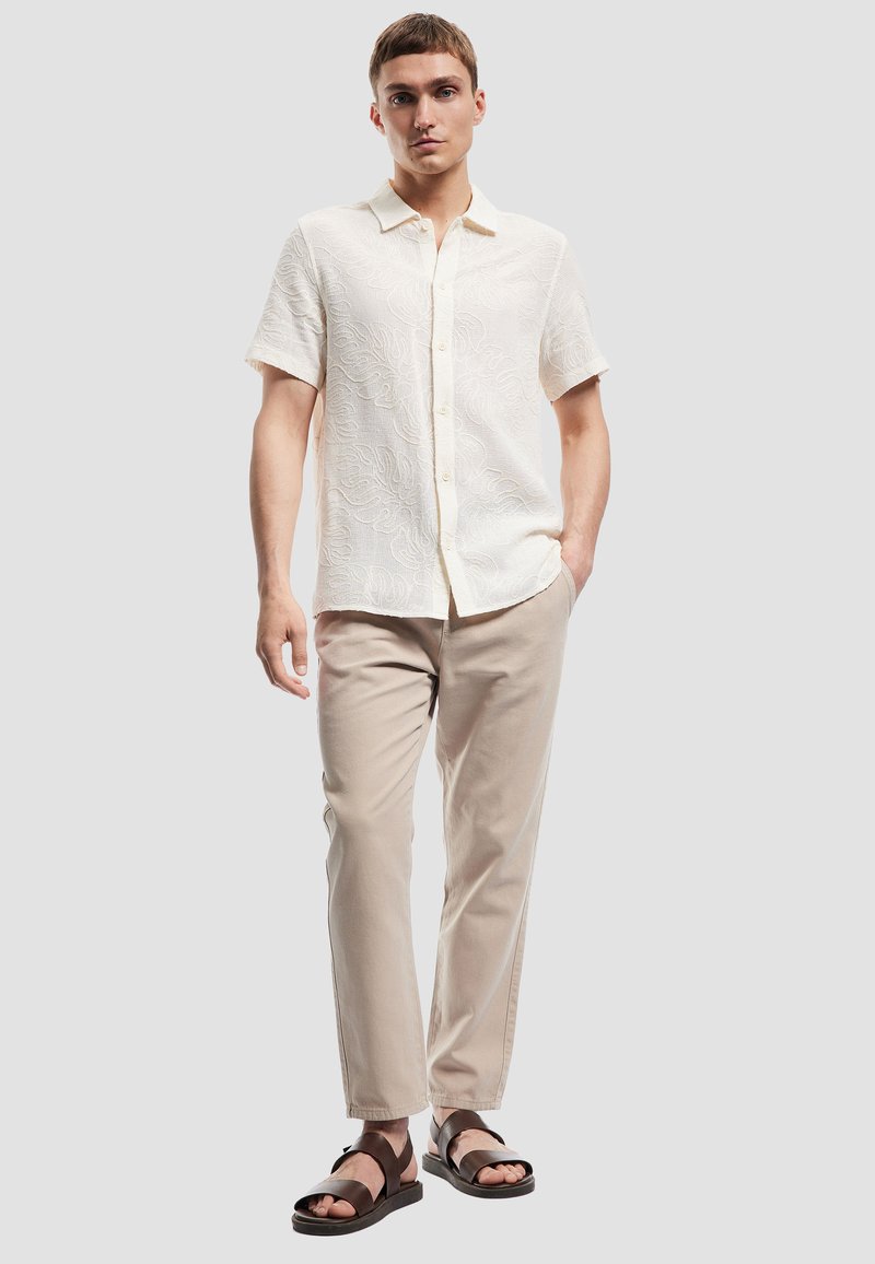 Korte mouw, crèmekleurig shirt met een textuurpatroon en knopen aan de voorkant, gecombineerd met lichtbeige broeken en zwarte sandalen.