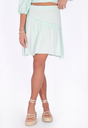 Donna che indossa una minigonna verde chiaro a balze con sandali beige con zeppa e laccio alla caviglia, in piedi con le gambe incrociate e le mani appoggiate sulla vita.