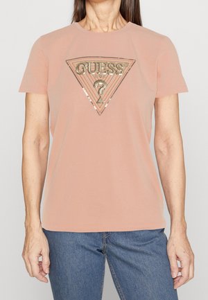 Kobieta ubrana w brzoskwiniowy T-shirt z krótkim rękawem, z cekinowym logo "GUESS?" w kształcie trójkąta, oraz niebieskie dżinsy.