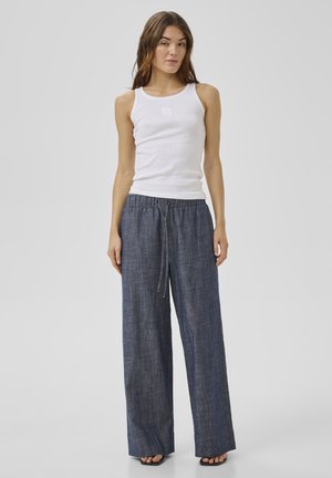 Kobieta ubrana w biały bezrękawnik i luźne, ciemnoniebieskie spodnie o szerokich nogawkach z paskiem ściągającym w talii, stojąca na jednolitym tle.