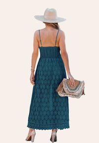 Teal maxi-jurk met een smocked lijfje, kantpatroon en een franjerugzak. Het model draagt een hoed met brede rand en sandalen met hak.