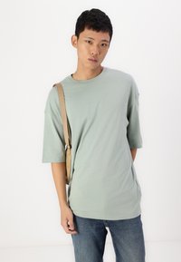 Světlá zelená oversize tričko s krátkými rukávy, kulatým výstřihem a jemnou, hladkou texturou, doplněné modrými džíny a světle hnědou křížovou taškou.